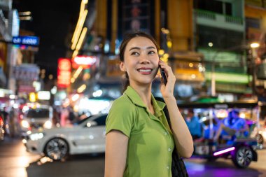 Asyalı çekici kadın sırt çantalı, şehirde yürürken telefonla konuşuyor. Güzel kadın turist gezgin akıllı telefon kullanarak tek başına seyahat ediyor. Tatilde gece yollarında iyi vakit geçiriyor.