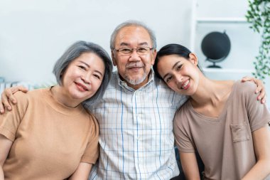 Evde kızıyla birlikte kanepede oturan Asyalı yaşlı çift portresi. Çekici güzel torun mutlu ve rahat hissediyor, olgun büyükbabayı ziyaret ediyor ve evde kameraya bakıyor..