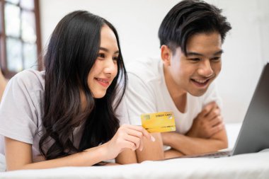 Asyalı genç çift evdeki yatak odasında dizüstü bilgisayar kullanıyor. Çekici, romantik, yeni evli bir erkek ve kadın yatakta uzanıp boş zamanlarını internet sitesinde alışveriş yaparak geçiriyorlar.