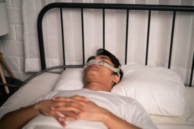 Cpap makinesi takan Asyalı adam bütün gece yatak odasında rahat uyuyacak. Çekici, yakışıklı, sağlıklı bir erkek, uyku apnesi oksijen maskesi teçhizatı kullanarak yatağa uzanırken mutlu ve rahat hisseder..