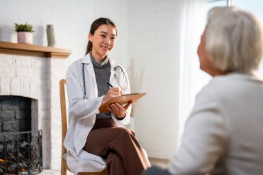 Asyalı genç bakıcı hemşireyi muayene et ve yaşlı kadın hastayı dinle. Çekici uzman bakıcı kadınlar, evdeki yaşlı olgun büyükannelere danışmanlık ve danışmanlık yaparlar..