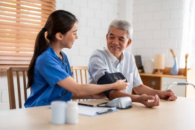 Asyalı bakıcı doktor yaşlı hastayı muayene ediyor. Kan basıncı göstergesi kullanıyor. Huzurevindeki kanepede otururken yaşlı bir adamla ilgilenen genç kadın terapist. Sağlık sigortası kavramı.