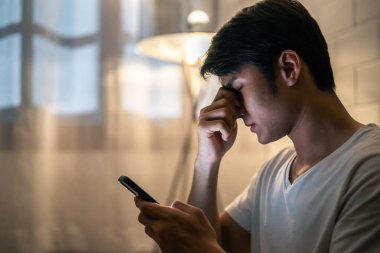 Yorgun Asyalı genç adam gece cep telefonu kullanırken gözünü yaraladı. Çekici erkek kanepede oturuyor, film izlemekten yorgun düşmüş göz yorgunluğu ve evde kuru göz masajı..