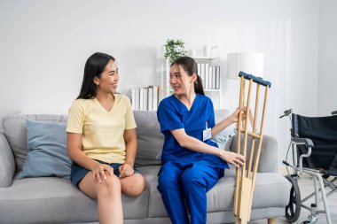 Asyalı kadın bacak ampütesi hemşirenin desteğiyle fizyoterapi yapıyor. Çekici genç kız bacaksız hasta oturma odasında kadın bakıcı doktorla yavaşça yürüme alıştırması yapıyor..