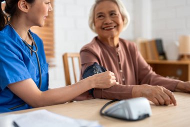 Asyalı bakıcı doktor yaşlı hastayı muayene ediyor. Kan basıncı göstergesi kullanıyor. Huzurevindeki kanepede otururken yaşlı kadınla ilgilenen genç kadın terapist. Sağlık sigortası kavramı.