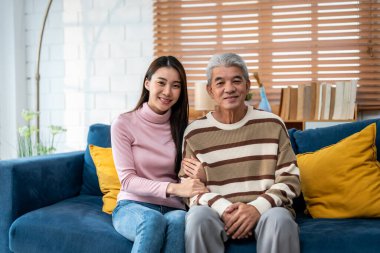 Evde kızıyla birlikte kanepede oturan Asyalı yaşlı babanın portresi. Çekici güzel torun mutlu ve rahat hissediyor, olgun büyükbabayı ziyaret ediyor ve evde kameraya bakıyor..