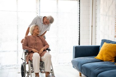 Asyalı aktif kıdemli adam, yaşlı kadını tekerlekli sandalyeden kanepeye kadar destekliyor. Mutlu bir aile, güzel olgun bir çift emeklilikten sonra evde birlikte oturma odasında fizik tedavi yapıyorlar..