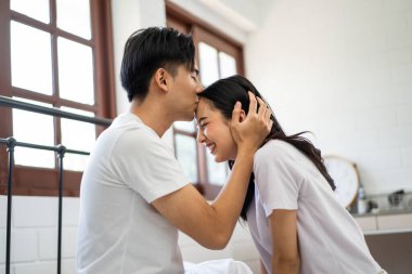 Asyalı bir eş, evdeki yatak odasında yatarken kocasını öpüyor. Pijamalı yeni evli çift mutlu ve rahatlamış. Sabahın erken saatlerinde evde birlikte vakit geçiriyorlar..