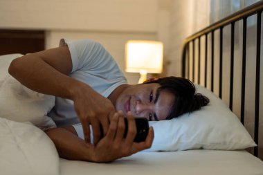 Asyalı genç yakışıklı adam gece yatak odasında cep telefonu kullanıyor. Çekici erkek, karanlık odada yatağa uzanmış, mutlu ve rahat bir şekilde film seyrediyor ve akıllı telefondan sohbet ediyor.