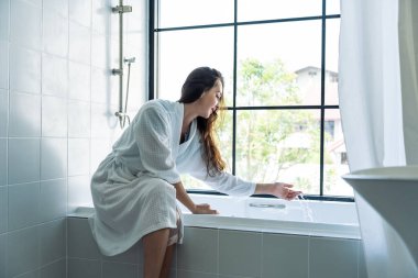 Beyaz kadın küvette oturmuş banyoda duş almak için su hazırlıyor. Bornozlu çekici kadın köpüğe ve suya dokunuyor ve sabahları sıcacık evdeki spa prosedürlerinin tadını çıkarıyor..