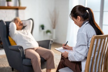 Asyalı bakıcı hemşire muayene ediyor ve yaşlı stresli kadın hastayı dinliyor. Çekici uzman bakıcı kadınlar, evdeki yaşlı olgun büyükannelere danışmanlık ve danışmanlık yaparlar.