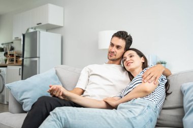 Beyaz, genç, sevgi dolu bir çift evdeki oturma odasında, kanepede oturuyor. Çekici yeni evli erkek ve kadın gülümsüyor ve birbirlerine sarılıyorlar. Sevgililer gününde evde birlikte vakit geçirmenin tadını çıkarıyorlar..