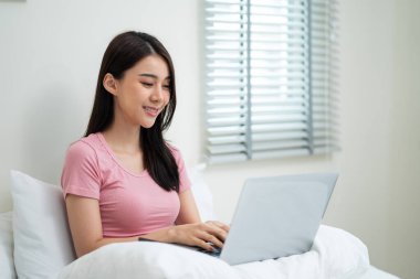 Güzel Asyalı genç iş kadını evde dizüstü bilgisayar klavyesi yazıyor. Çekici kız öğrenci çalışan yatakta oturur ve bilgisayar planlama projesini kullanır ve evdeki oturma odasında internetten öğrenir.