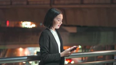 Asyalı genç iş kadını geceleri şehir merkezinde cep telefonuyla dışarıda duruyor. Akıllı telefon kullanırken ve modern ağ teknolojisiyle eğlenirken mutlulukla gülümseyen güzel kız..