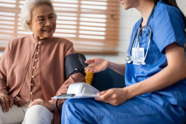 Asyalı bakıcı doktor yaşlı hastayı muayene ediyor. Kan basıncı göstergesi kullanıyor. Huzurevindeki kanepede otururken yaşlı kadınla ilgilenen genç kadın terapist. Sağlık sigortası kavramı.