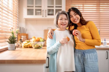 Evde kızıyla birlikte duran Asyalı yaşlı anne portresi. Çekici torun ve büyükanne mutlu ve rahatlamış hissediyorlar, sağlıklı meyveler tutuyorlar ve evdeki kameraya bakıyorlar..