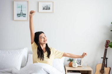 Asyalı genç ve güzel bir kadın sabah yataktan kalkıyor. Pijamalı çekici kız mutlu ve rahat hissediyor, uyanıyor ve hafta sonu yatak odasında vücudunu esnetiyor..