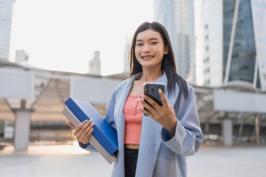 Şehirdeyken cep telefonu kullanan Asyalı iş kadınının portresi. Çekici genç kız kameraya bakıyor, elinde dijital akıllı telefon ekipmanları, gökdelen binası ve mutluluk dolu..