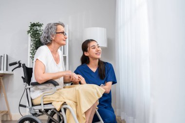 Beyaz, son sınıf öğrencisi kadın, genç Asyalı hemşireyle birlikte tekerlekli sandalyede oturuyor. Çekici kadın bakıcı doktor ziyareti, teşhis koymak ve evdeki oturma odasındaki yaşlı hastayı muayene etmek.