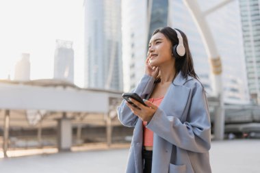 Şehirde kulaklık takan ve akıllı telefon kullanan Asyalı bir kadın. Çekici genç kız dijital cep telefonu tutuyor, açık havada müzik dinlemenin keyfini çıkarıyor, mutlulukla gökdelen inşa ediyor..