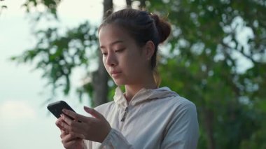 Spor kıyafetleri içinde Asyalı genç bir kadın parkta yürüyüş yaptıktan sonra arkadaşlarıyla cep telefonuyla iletişim kuruyor. Aktif güzel kız hafta sonu aktivitelerini arka bahçede yapıyor..