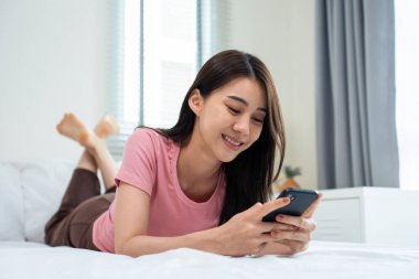 Asyalı çekici kadın sabah evde cep telefonu sohbetini kullanıyor. Mutlu, sıradan, güzel bir kadın yatak odasında uzanıyor. Tatil haftasonunun tadını çıkarın. Evdeki teknolojiyle akıllı telefon iletişimi kurun..