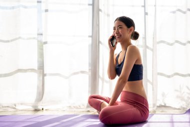 Evde yoga pilatesini yaparken Asyalı sporcu kadın telefonda konuşuyor. Akıllı telefon kullanarak internet üzerinden iletişim kuran ve egzersiz yapan çekici bir kız fitness yoga stüdyosunda sağlık için meditasyon yapıyor..