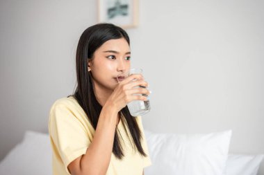 Asian attractive woman sit on bed, drink a glass of water in bedroom. Young thirsty beautiful girl holding clean mineral natural in cup after wake up and sipping in morning for health care in house.