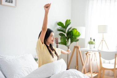 Asyalı genç ve güzel bir kadın sabah yataktan kalkıyor. Pijamalı çekici kız mutlu ve rahat hissediyor, uyanıyor ve hafta sonu yatak odasında vücudunu esnetiyor..