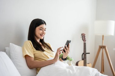 Asyalı çekici kadın sabah evde cep telefonu kullanıyor. Mutlu, sıradan, güzel bir kadın, yatak odasında oturmuş, akıllı telefondan konuşuyor teknolojiyle iletişim kuruyor, tatilin tadını çıkarıyor..