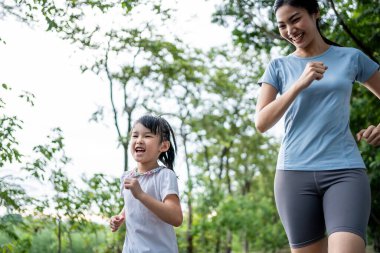 Asyalı genç anne ve kız bahçede koşuyorlar. Mutlu bir aile, spor giyim sektöründe çekici bir anne kendini mutlu hissediyor ve halka açık bir parkta sağlık için sevimli bir kızla egzersiz yapıyor.