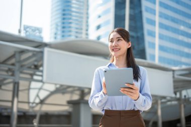 Asyalı genç iş kadını şehirde dikilirken dijital tablet kullanıyor. Yüksek binalarda, gökdelenlerde, açık havada teknolojiyle çalışan çekici bir kadın işçi.