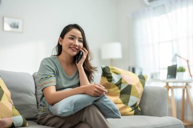Asyalı güzel kadın kanepede oturur ve cep telefonuyla konuşur. Mutlu çekici genç kız evde vakit geçirir, rahatlar, iletişim kurar ve oturma odasında akıllı telefondan tartışır..