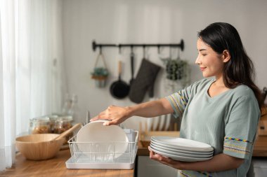 Asian active cleaning service woman worker cleaning in kitchen at home. Beautiful young girl housekeeper cleaner feel happy and wipes dishes plates after washing for housekeeping housework or chores.