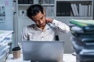 Yorgun Asyalı genç iş adamı gece geç saatlere kadar ofiste çalışıyor. Pijamalı çekici adam yorgun ve bitkin hissediyor, iş yerinde dizüstü bilgisayar planı projesi kullanırken boynuna masaj yapıyor..