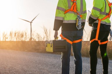 Rüzgar türbinleri tarlasında yürüyen iki teknisyene yakın durun. Yenilik mühendisi güvenlik donanımı giyen adamlar, çevresel yenilenebilir temiz enerji geliştirmek için yel değirmeni çiftliğinde çalışıyorlar.