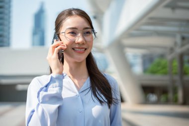 Asyalı genç iş kadını şehirde dikilirken akıllı telefon kullanıyor. Çekici, güzel bir bayan çalışan cep telefonuyla iş arkadaşlarıyla açık havada güven içinde iletişim kuruyor..
