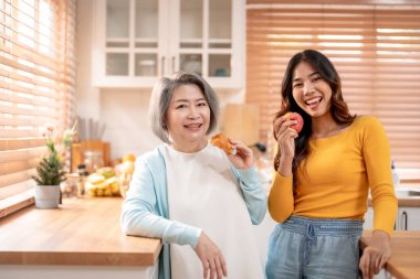 Evde kızıyla birlikte duran Asyalı yaşlı anne portresi. Çekici torun ve büyükanne mutlu ve rahatlamış hissediyorlar, sağlıklı meyveler tutuyorlar ve evdeki kameraya bakıyorlar..