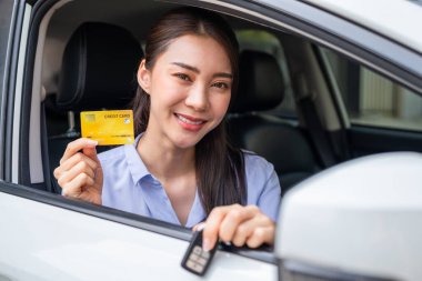Arabada otururken kredi kartı tutan Asyalı kadın sürücünün portresi. Çekici, mutlu, genç kız müşteri arabanın içinden kameraya bakıyor. Aracın içindeki alışveriş kartını özgüvenle gösteriyor..