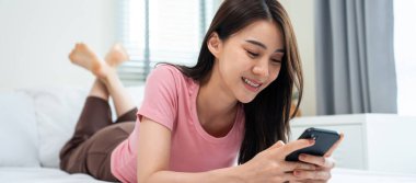 Asyalı çekici kadın sabah evde cep telefonu sohbetini kullanıyor. Mutlu, sıradan, güzel bir kadın yatak odasında uzanıyor. Tatil haftasonunun tadını çıkarın. Evdeki teknolojiyle akıllı telefon iletişimi kurun..