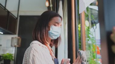 Çekici garson, kafede Covid-19 salgınını önlemek için koruyucu maske takıyor. Genç işletme sahibi kız barista apron temiz ayna ile dezenfektan ve hoş geldiniz müşteri önce silme