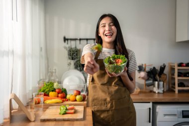 Asyalı kadın portresi evde mutfakta yeşil sağlıklı salata pişiriyor. Önlük giyen güzel bir genç kız, sabahları evdeki sağlık hizmetleri için temiz yemek yapmaktan zevk alıyor ve gülümsüyor, kameraya bakıyor..