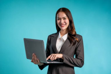 İzole edilmiş mavi arka planda duran Asyalı iş kadını portresi. Resmi olarak giyinmiş çekici genç bayan çalışanlar dizüstü bilgisayar kullanıyor ve gülümsüyor. Stüdyo ekranındaki kameraya bakın..