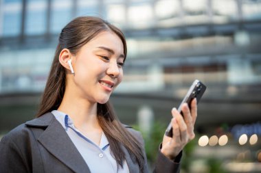 Asyalı iş kadını şehirde dururken kulaklıkla müzik dinler. Çekici kadın çalışan kendini mutlu ve rahat hissediyor. Dışarıda cep telefonuna bakıyor ve iş yerinde dinleniyor.