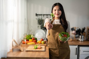 Asyalı kadın portresi evde mutfakta yeşil sağlıklı salata pişiriyor. Önlük giyen güzel bir genç kız, sabahları evdeki sağlık hizmetleri için temiz yemek yapmaktan zevk alıyor ve gülümsüyor, kameraya bakıyor..