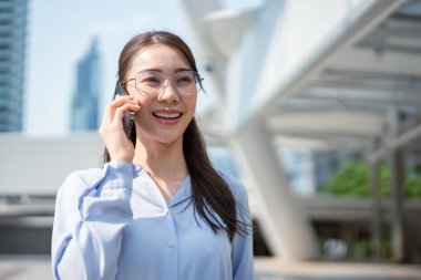 Asyalı genç iş kadını şehirde dikilirken akıllı telefon kullanıyor. Çekici, güzel bir bayan çalışan cep telefonuyla iş arkadaşlarıyla açık havada güven içinde iletişim kuruyor..