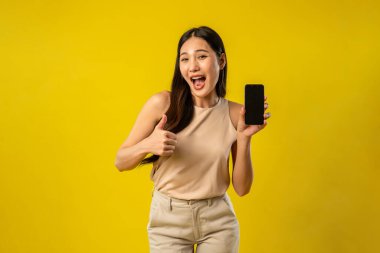 İzole edilmiş sarı arka planda akıllı telefonu tutan Asyalı kadının portresi. Çekici genç kız manken stüdyo ekranında çevrimiçi dijital cep telefonu aygıtı kullanırken mutlu ve neşeli hisseder..