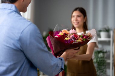 Asyalı genç çiçekçi kız dükkandaki müşteriye güzel buket satıyor. Çekici bayan küçük işletme sahibi kendini mutlu hissediyor ve rahatlıyor. Çiçek hediye paketini çiçekçi dükkanındaki erkek alıcıya veriyor.