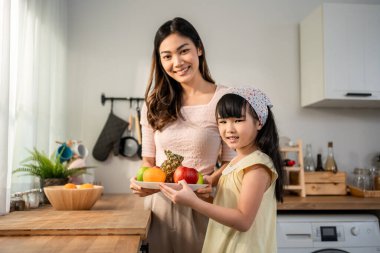 Asyalı anne ve genç kızın sağlıklı meyve tutarken portresi. Annesinin yanında duran sevimli çocuk, evde sağlıklı beslenme ve sağlık için sağlıklı gıdalar yemekten ve kameraya bakmaktan zevk alıyor..