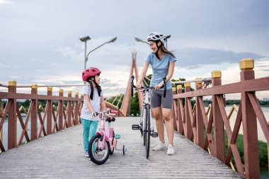 Asyalı anne ve kızı köprüde bisikleti tepeye itiyorlar. Mutlu bir aile, spor giyim sektöründe çekici bir anne kendini mutlu hissediyor ve sağlık hizmetleri için sevimli bir kızla parkta egzersiz yapıyor.